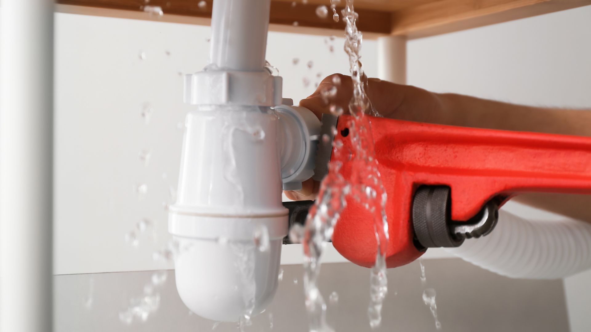 Hand using a red wrench on a leaking white sink pipe. Water sprays.