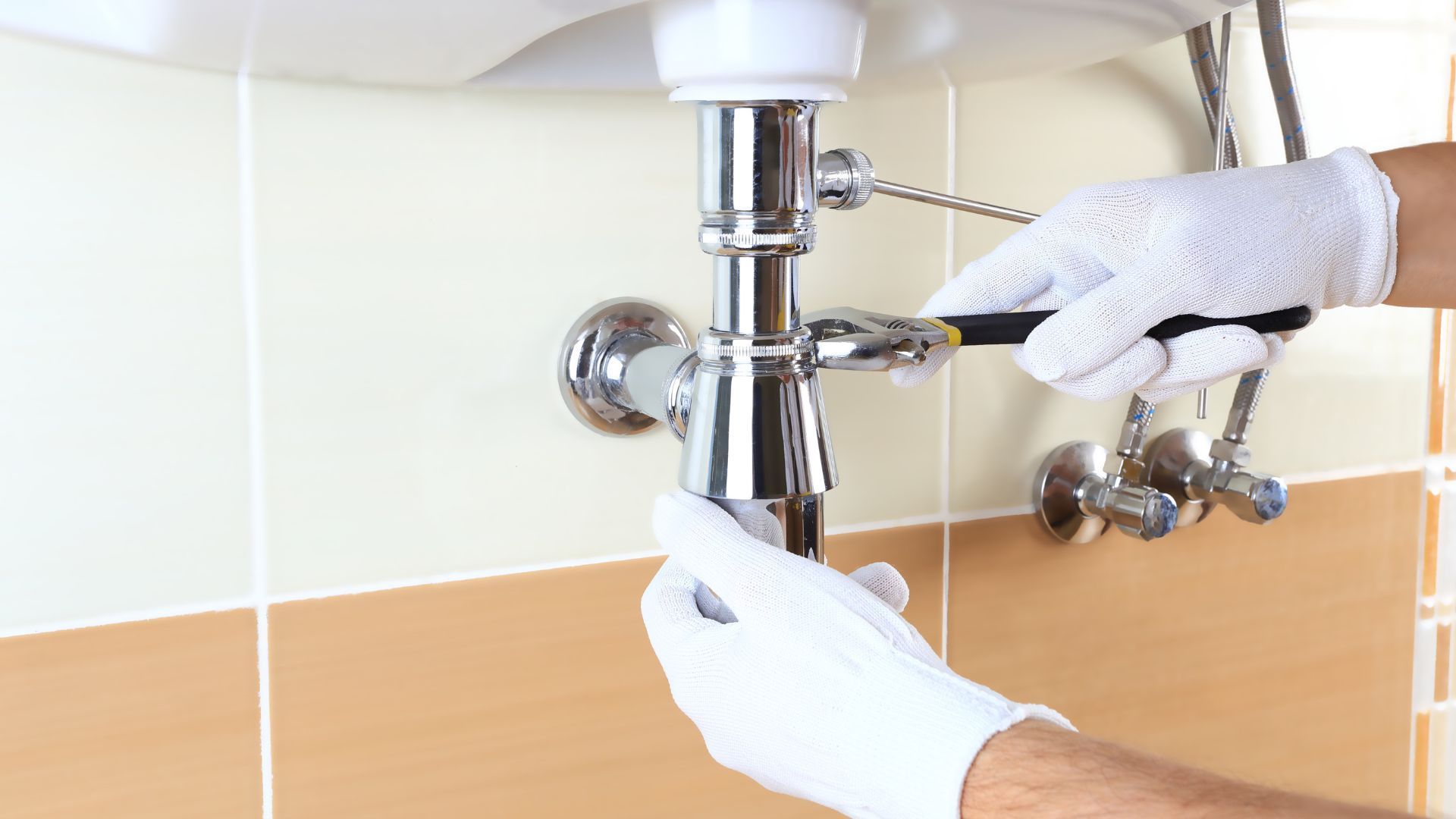 Plumber in white gloves working on a chrome sink drain with a wrench in a tiled bathroom.