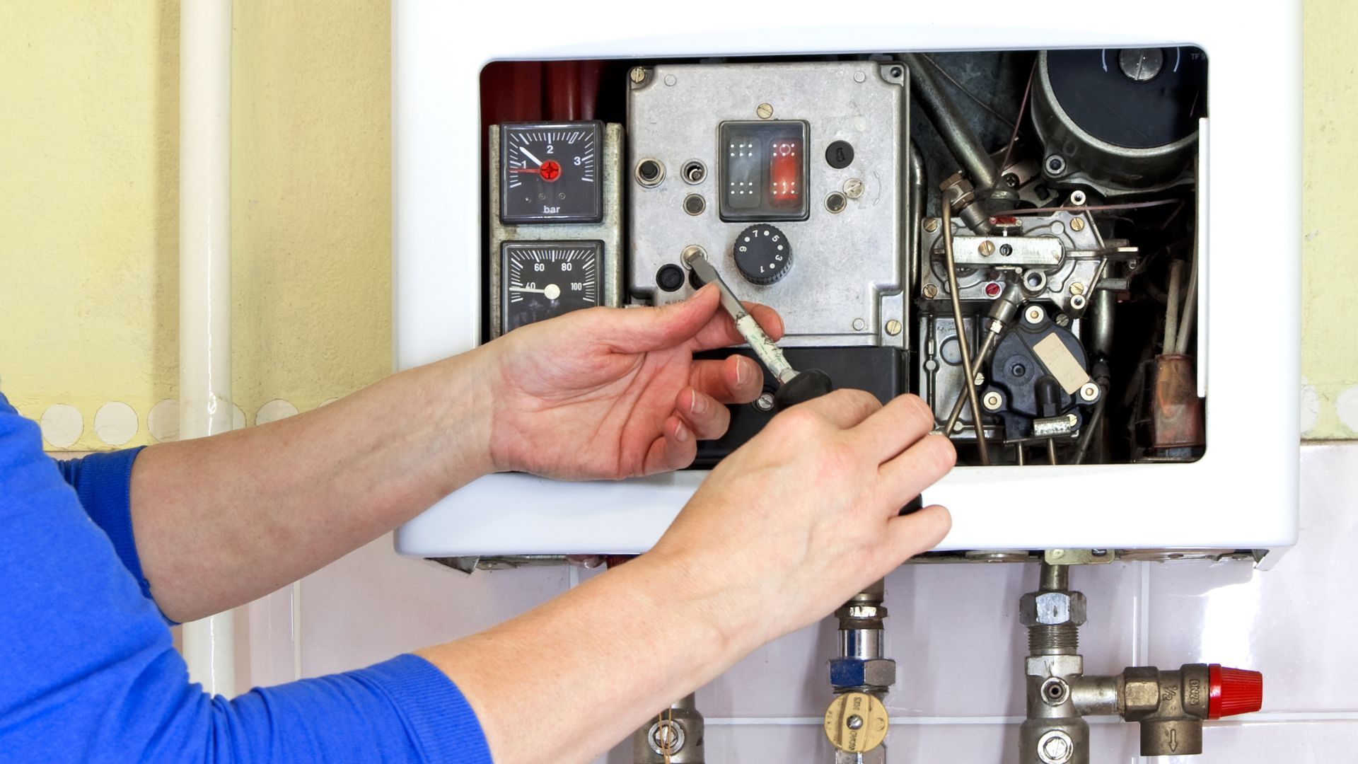 Person using a screwdriver to repair a boiler. Blue shirt, indoor setting, white boiler.