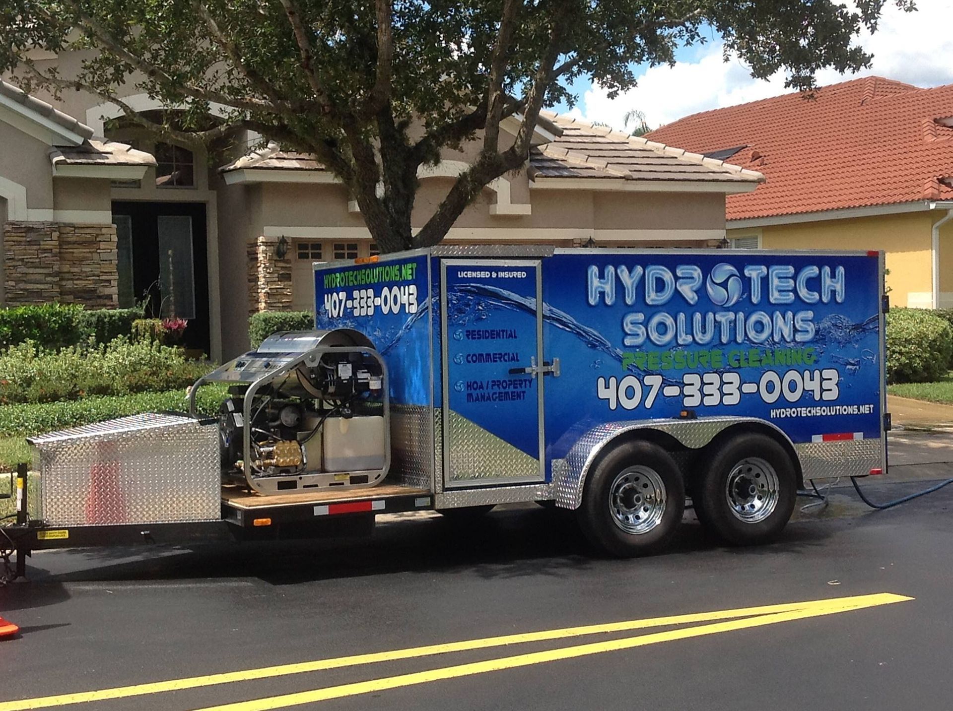A Hydrotech Solutions trailer parked on a street in front of a house, featuring blue branding and a visible engine.
