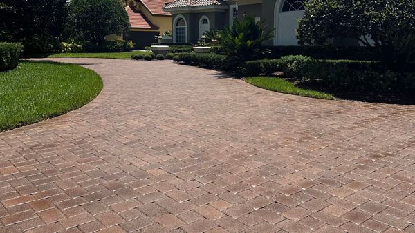 A curved driveway paved with multi-toned red and brown brick pavers, leading toward a house and landscaped garden.