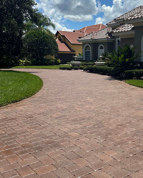 Brick driveway curves toward a house with red tile roof and manicured green lawn under a partly cloudy sky