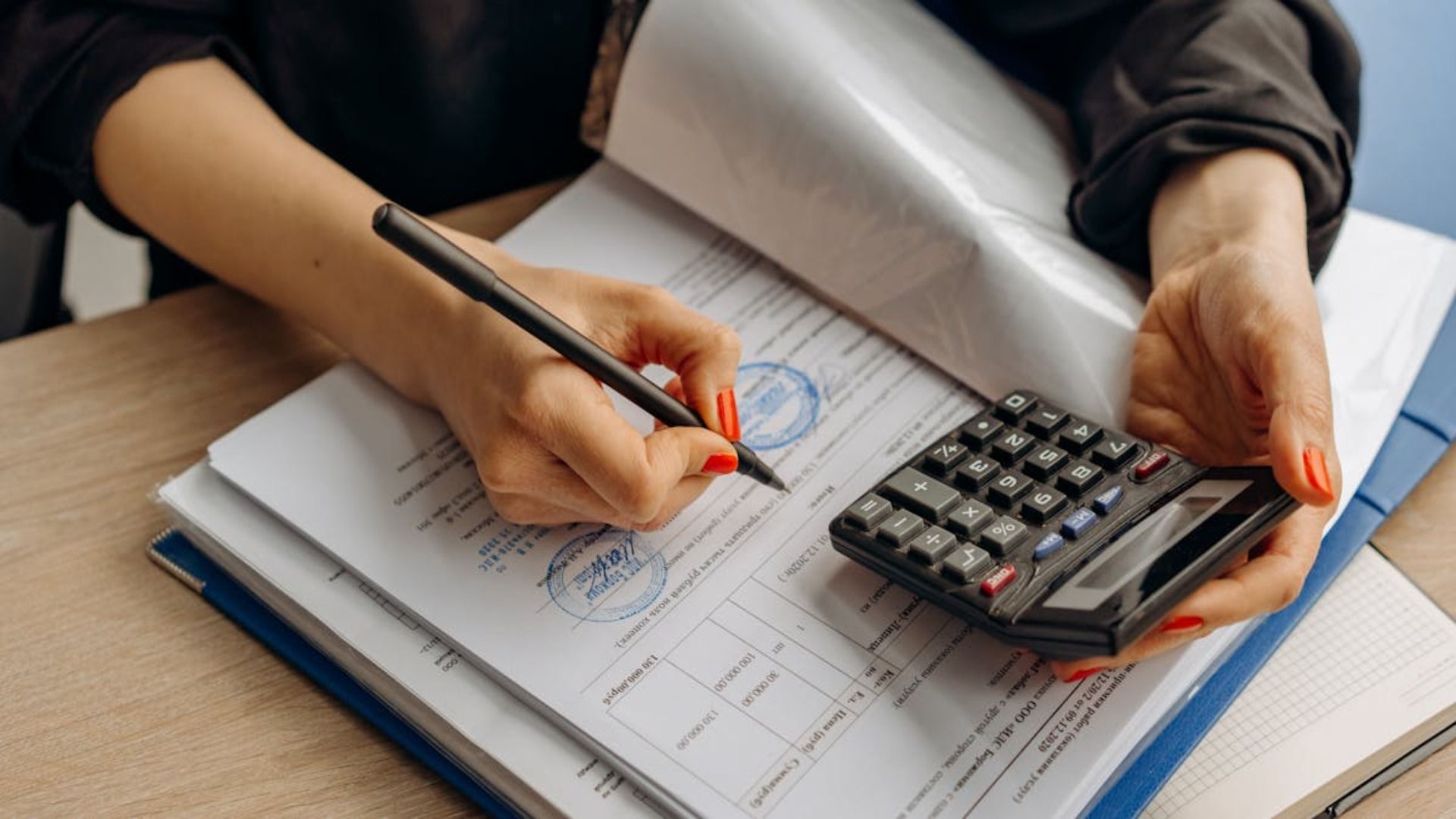 Unas manos sostienen un bolígrafo y una calculadora mientras revisan documentos financieros en un escritorio de madera.
