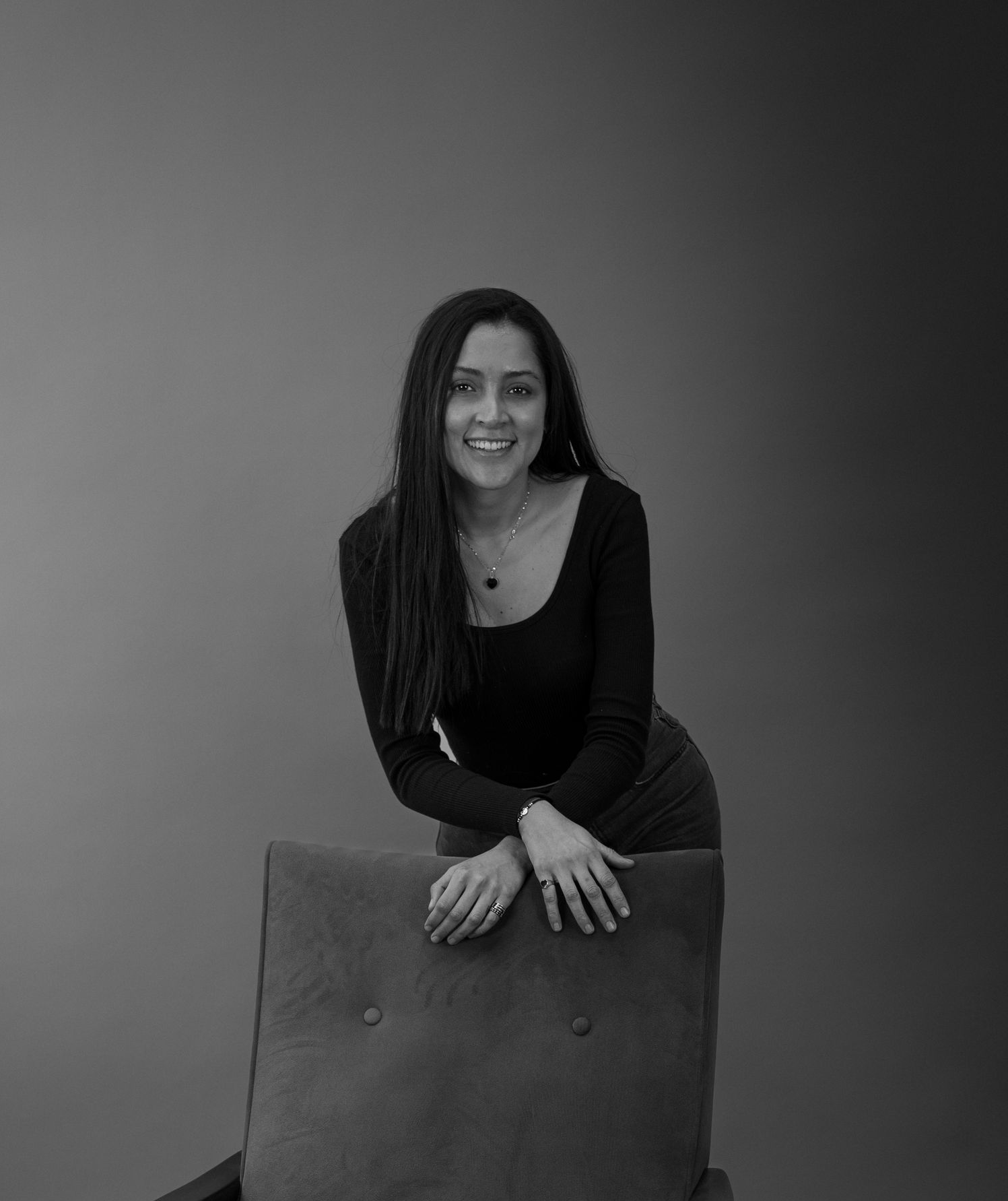 Woman in a black top smiling and leaning on a chair against a plain gray backdrop — Gracia Building Design In Kiama, NSW