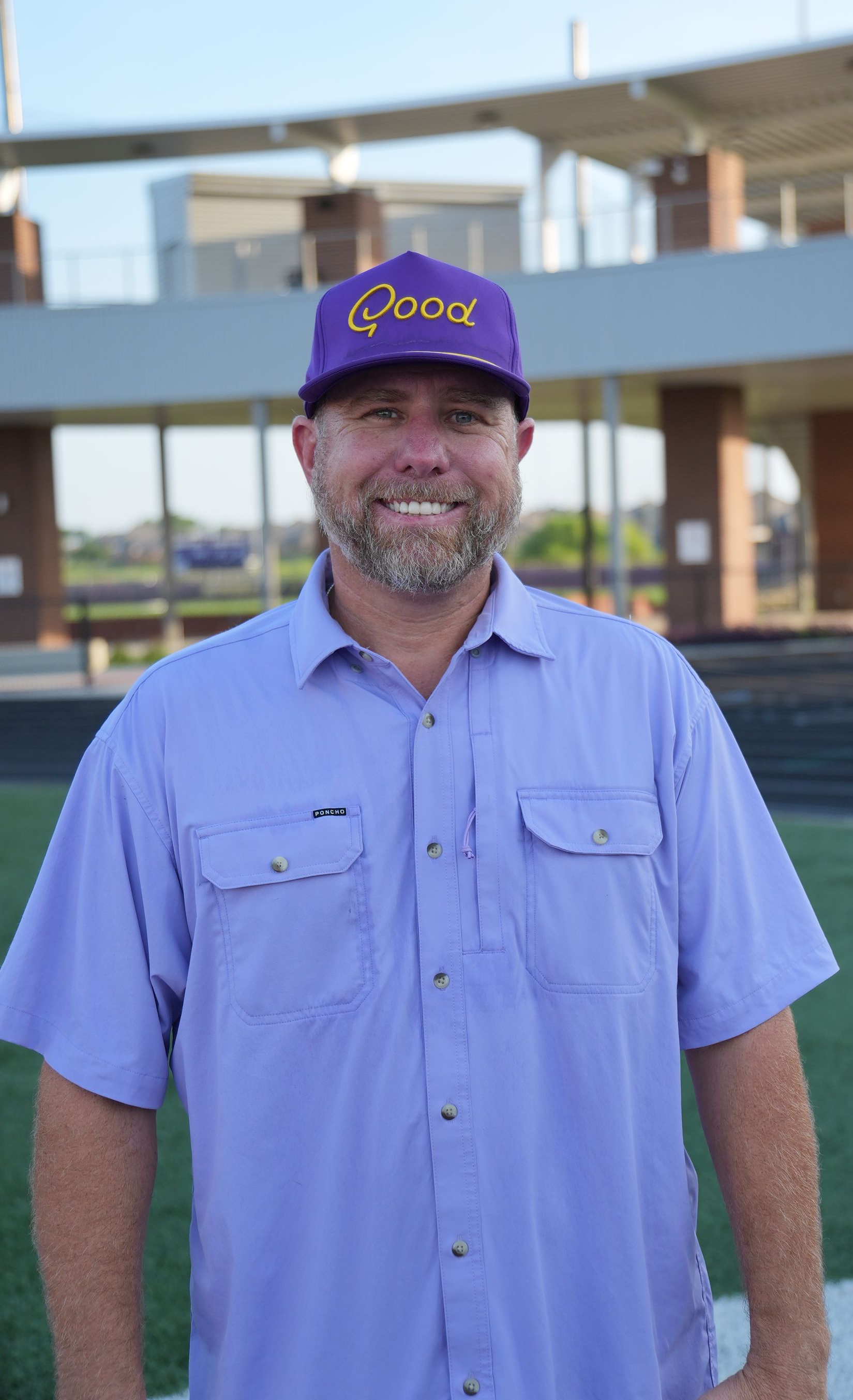 Football coach, arms crossed, on the field. Blue shirt, serious expression, players in background.