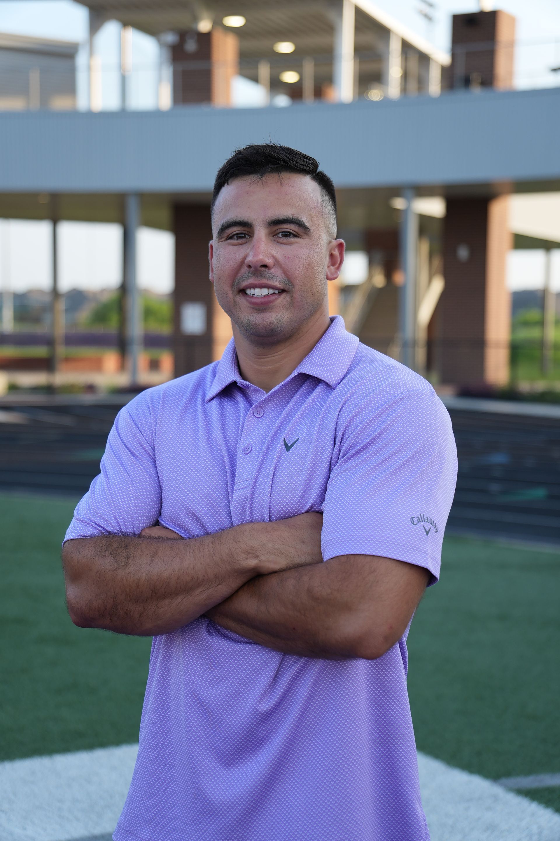 Football coach, arms crossed, on the field. Blue shirt, serious expression, players in background.