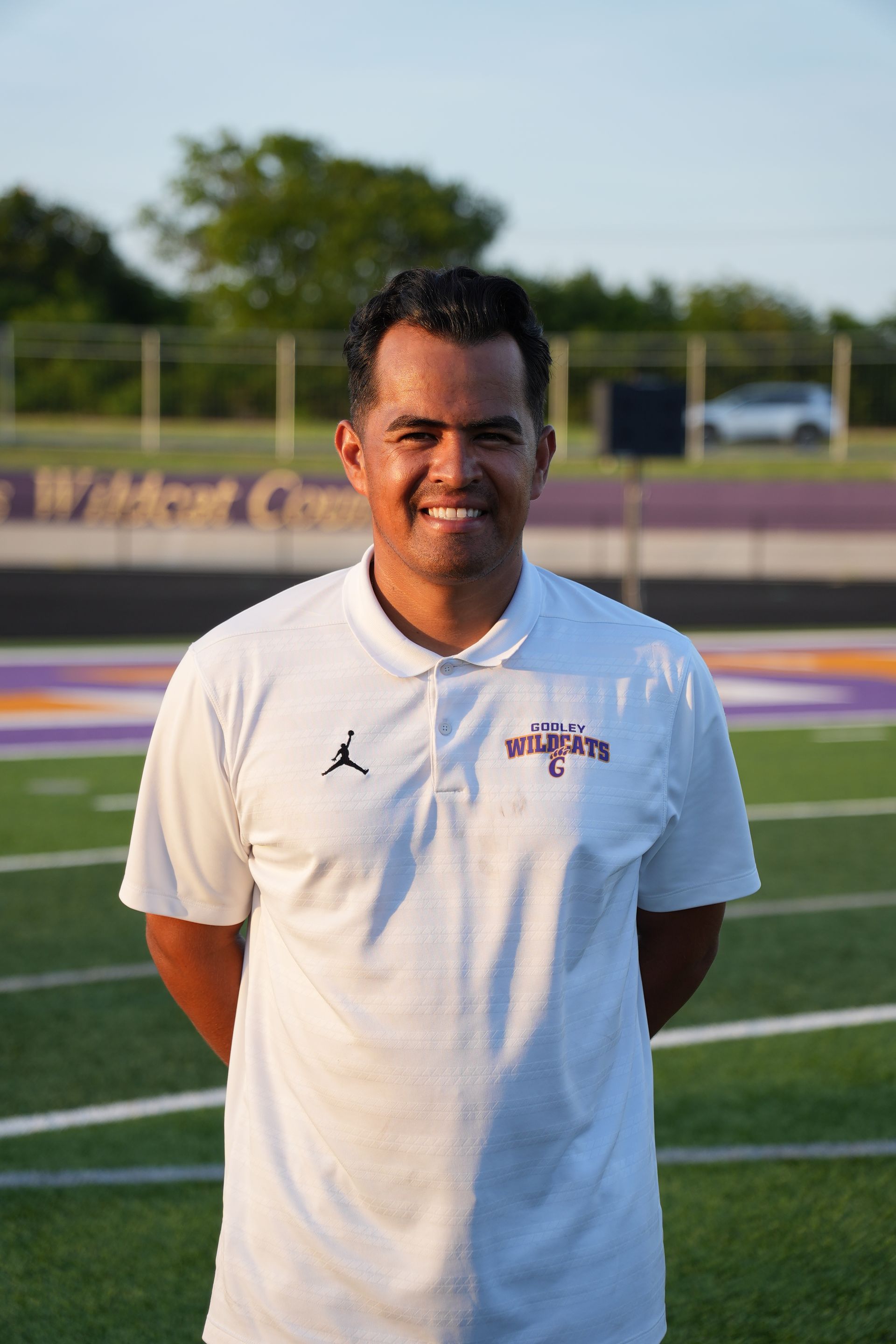 Man in green polo stands on a football field, arms crossed, stadium in background.