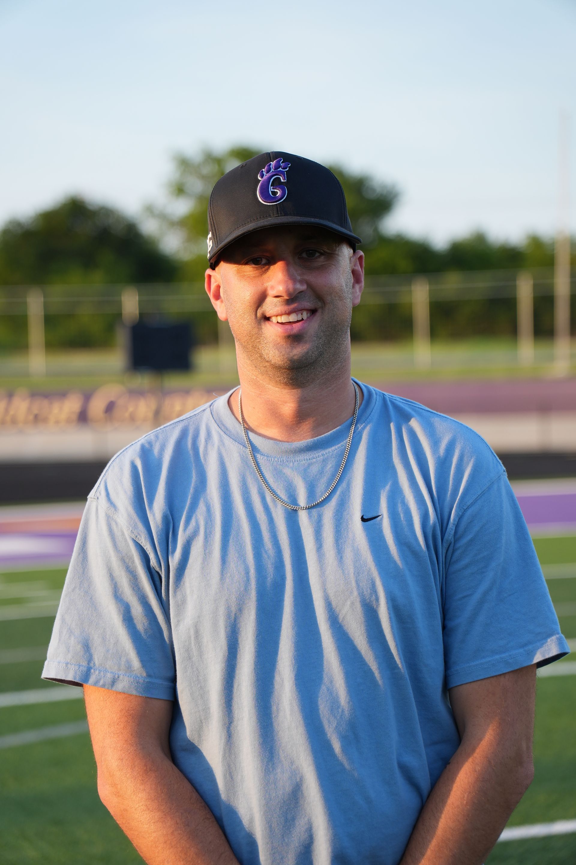 Man in green polo stands on a football field, arms crossed, stadium in background.