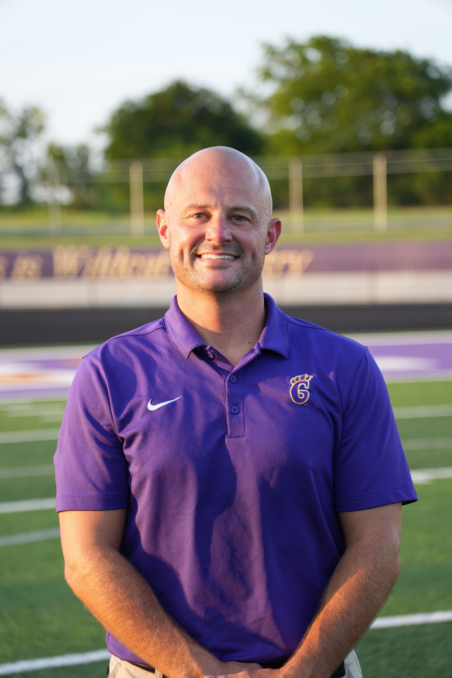 Man with crossed arms on a football field. He is wearing a blue polo shirt, khaki pants, and has a serious expression.