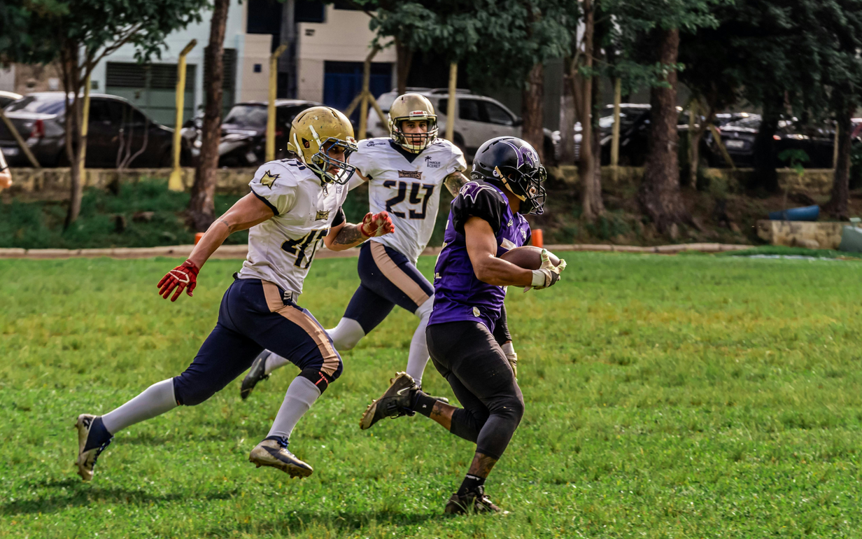 Football players in action on a grassy field; one runs with the ball, pursued by two opponents.
