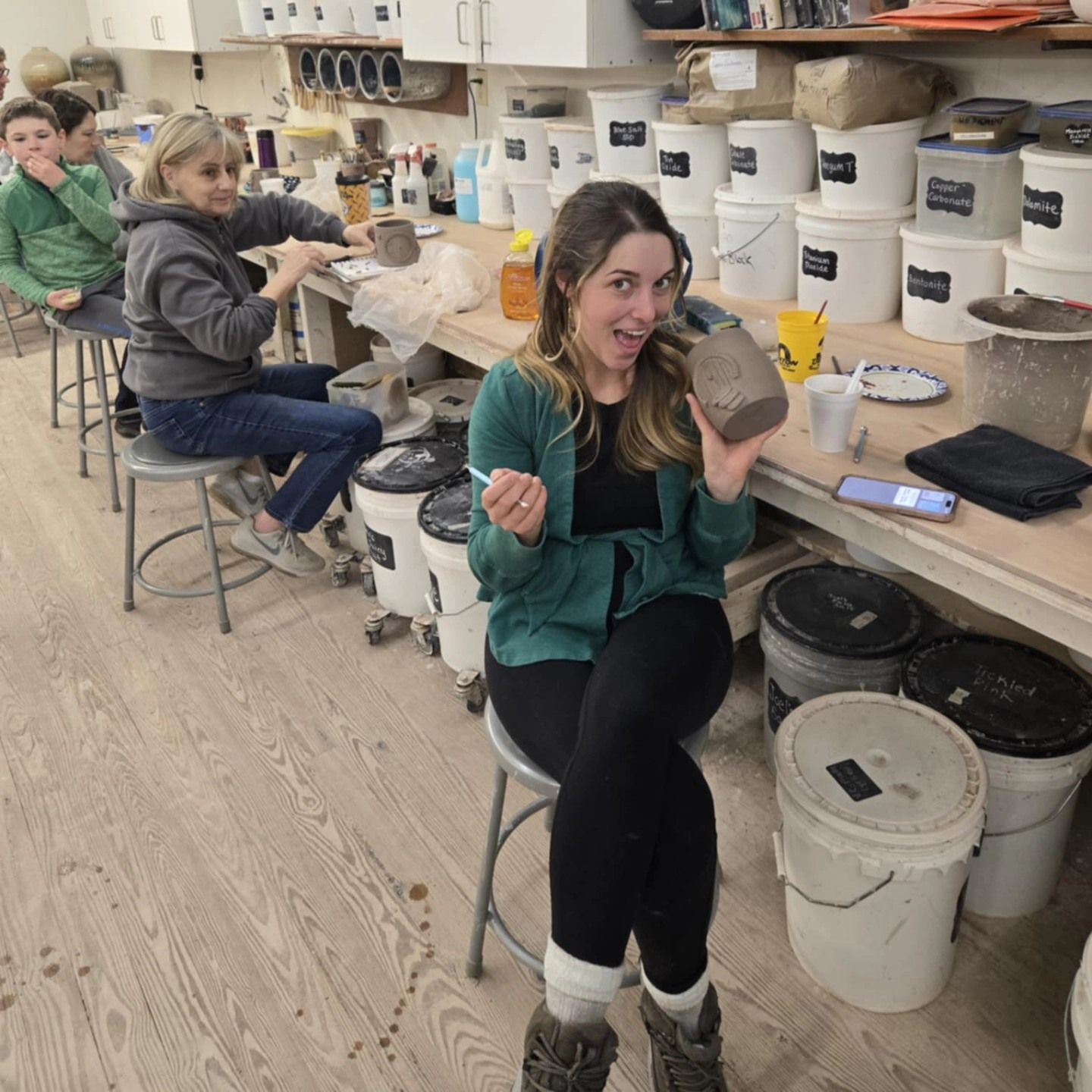 Woman in pottery studio holding clay, smiling. Students in background, white buckets, wood floor.