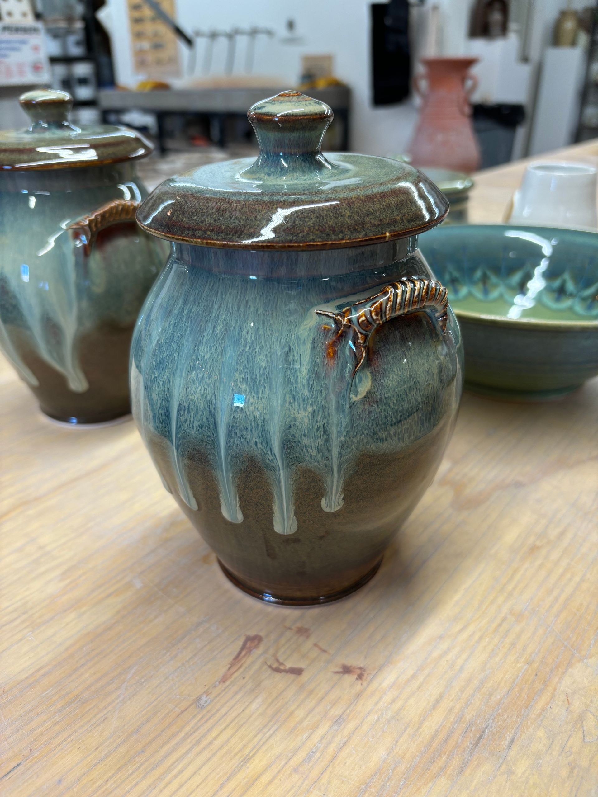 A glazed ceramic jar with lid, teal and brown with dripping glaze, sitting on a wooden surface.