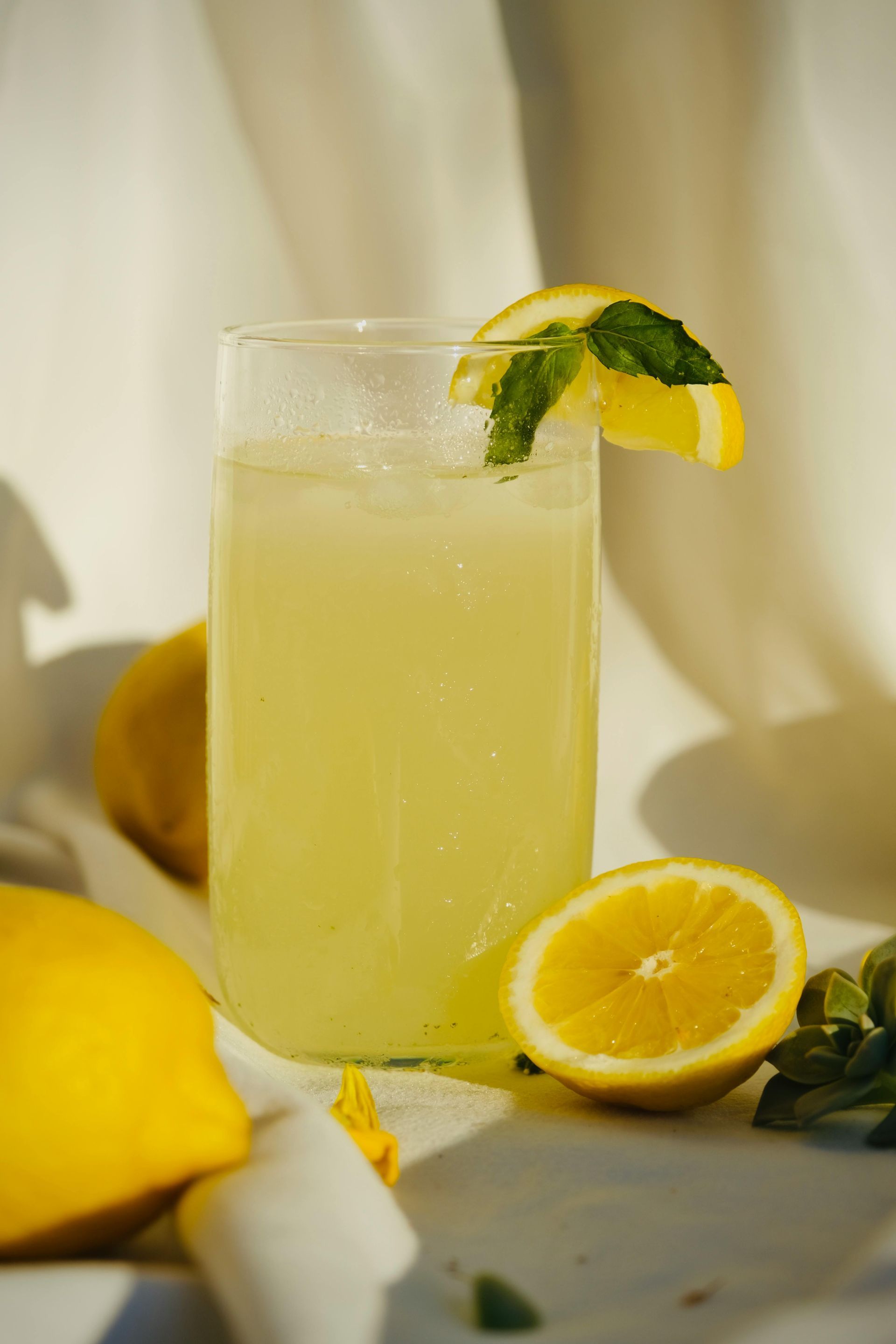 Tall glass of pale yellow lemonade with mint and lemon garnish, surrounded by fresh lemons on a table