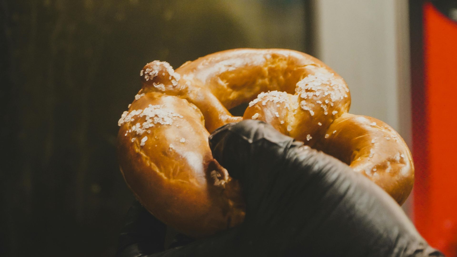 A gloved hand holding a glossy pretzel with coarse salt, lit by warm orange light.