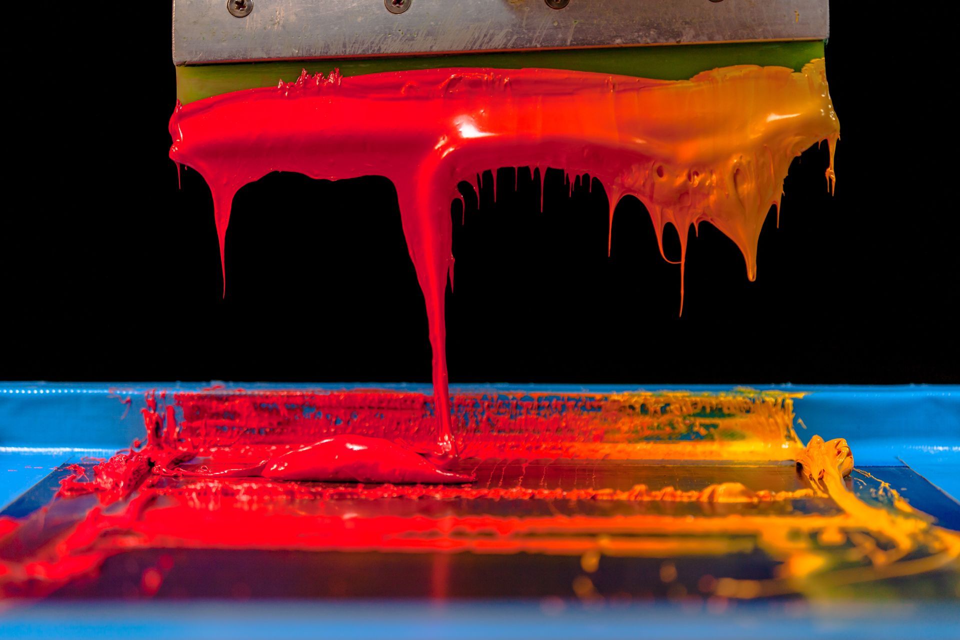 Colorful paint dripping from a squeegee onto a blue surface, blending red, pink, and yellow.
