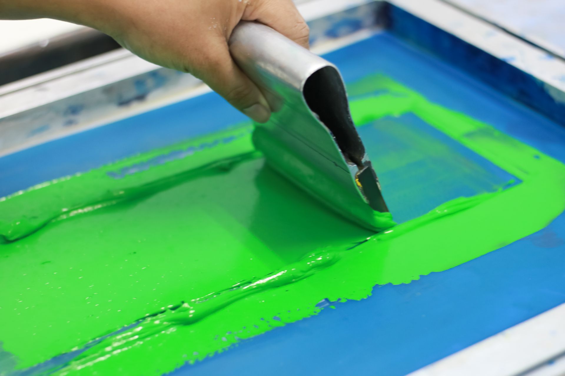 Hand spreading bright green paint through a blue-framed screen print stencil