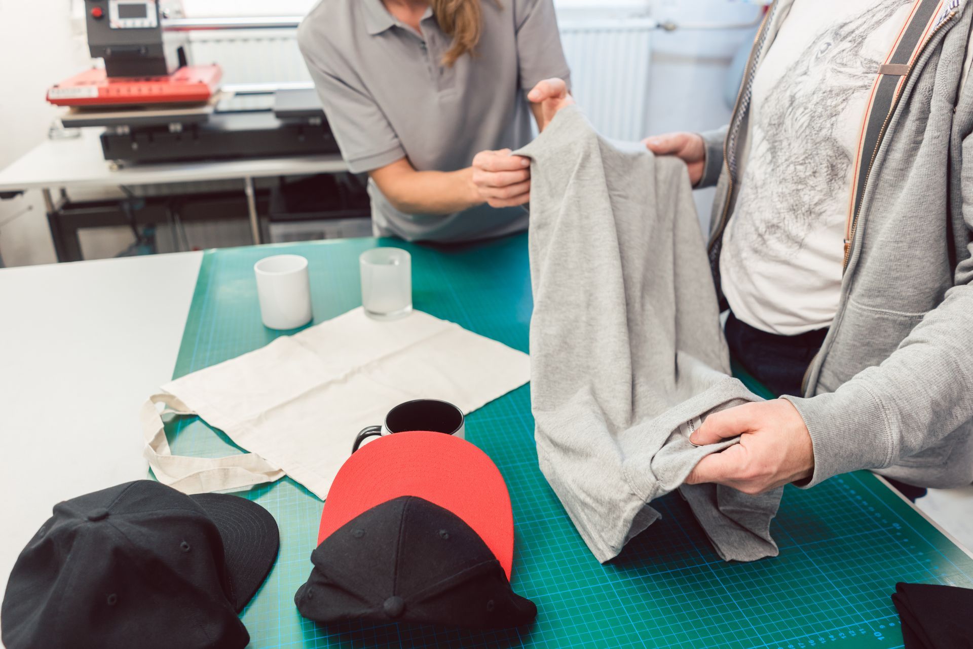 People folding a gray shirt at a worktable with fabric samples and hats