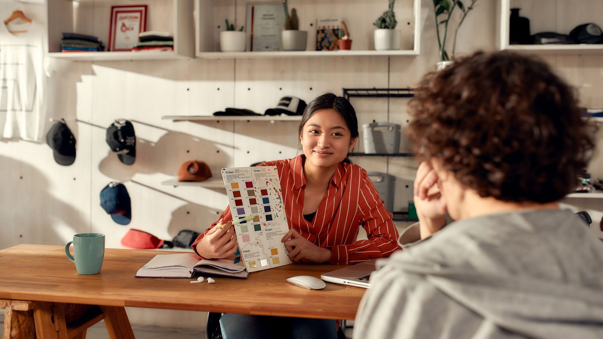 Two people talking at a kitchen table, one holding a colorful notebook in a bright home office.