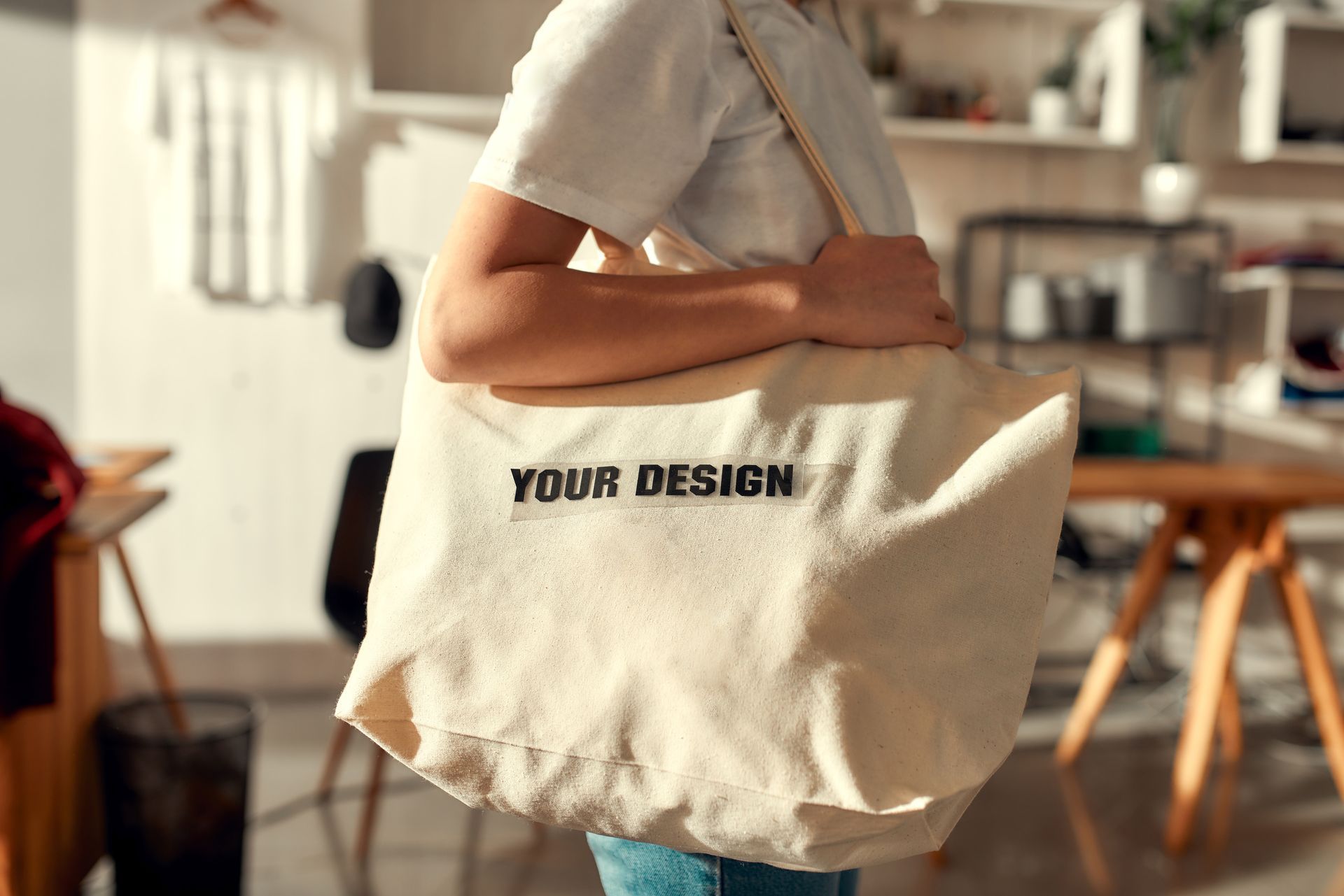 Person carrying a beige tote bag printed with “YOUR DESIGN” in a bright kitchen