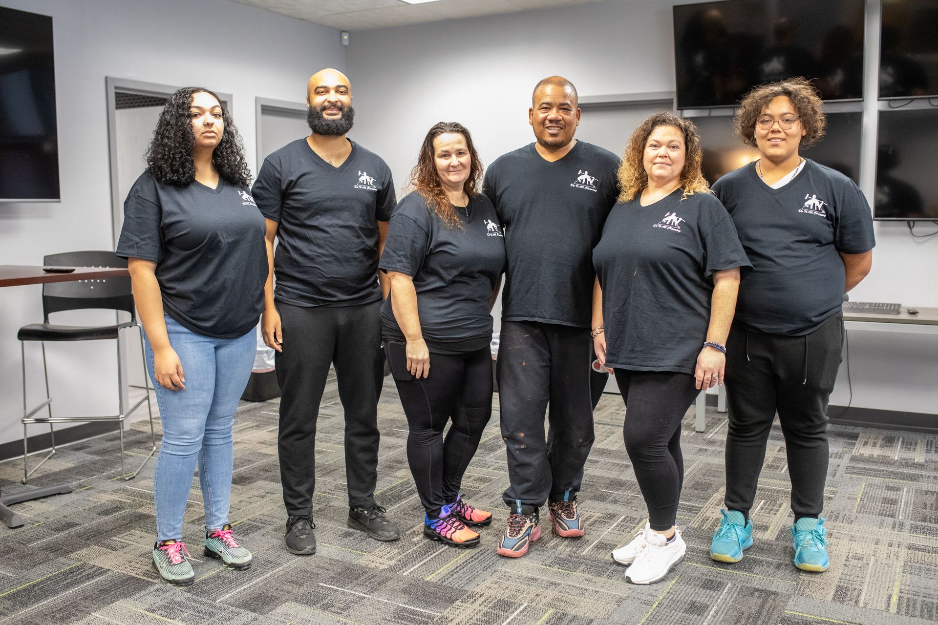 Six people in black shirts pose indoors, smiling. They stand in a room with monitors and neutral walls.