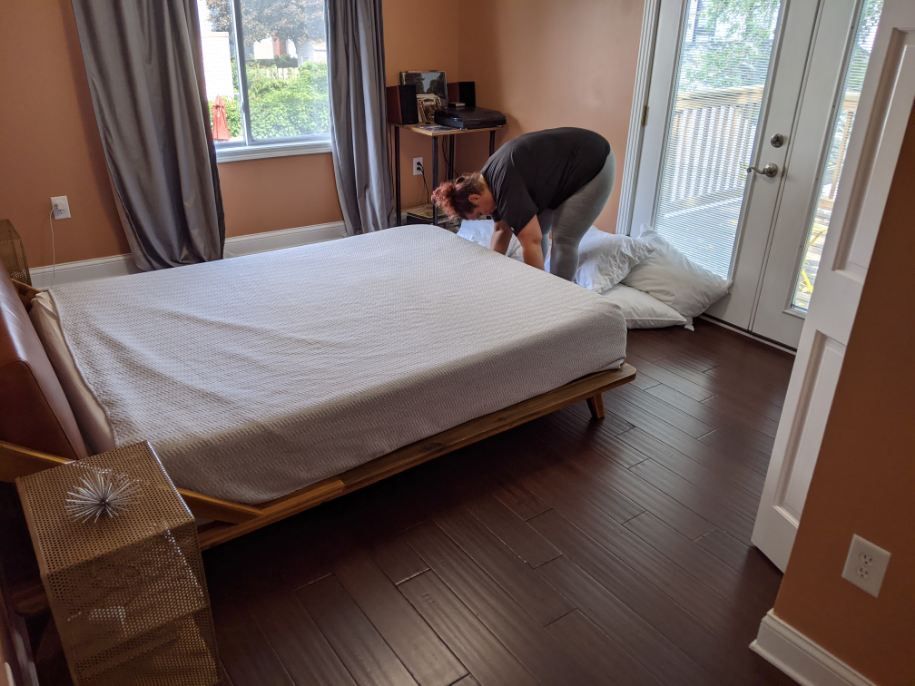 Person making a bed in a room with hardwood floors, a window, and a door leading to a balcony.
