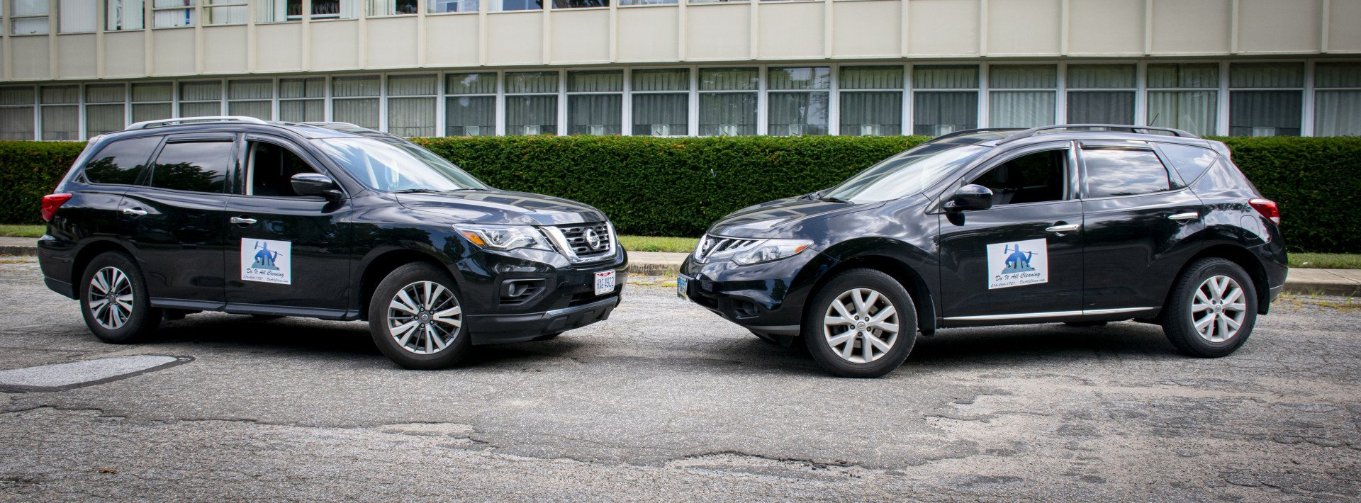 Two black SUVs parked side-by-side on a gray road in front of a building.