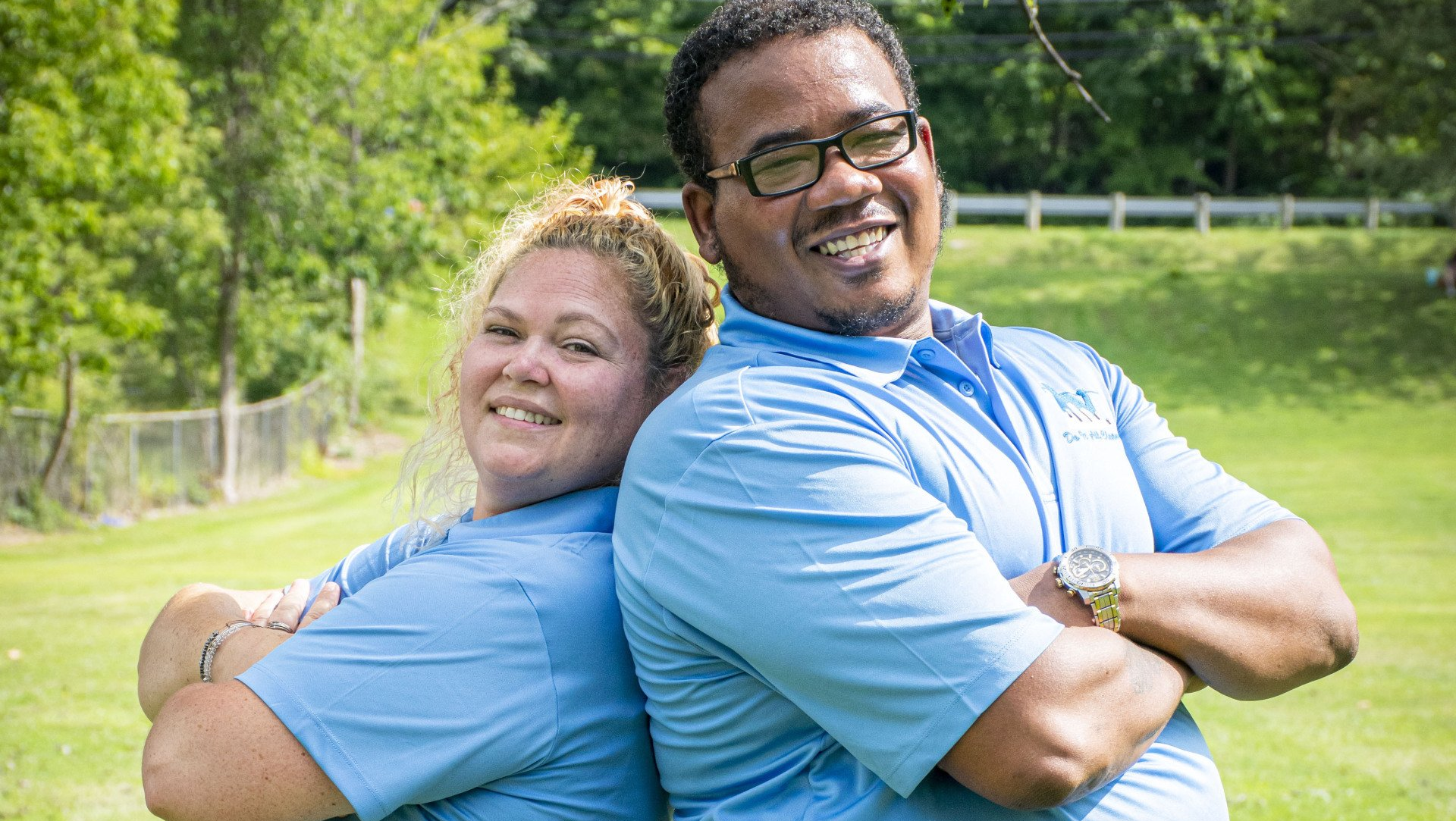 Two people in blue shirts stand back-to-back, smiling, outdoors on a grassy lawn.
