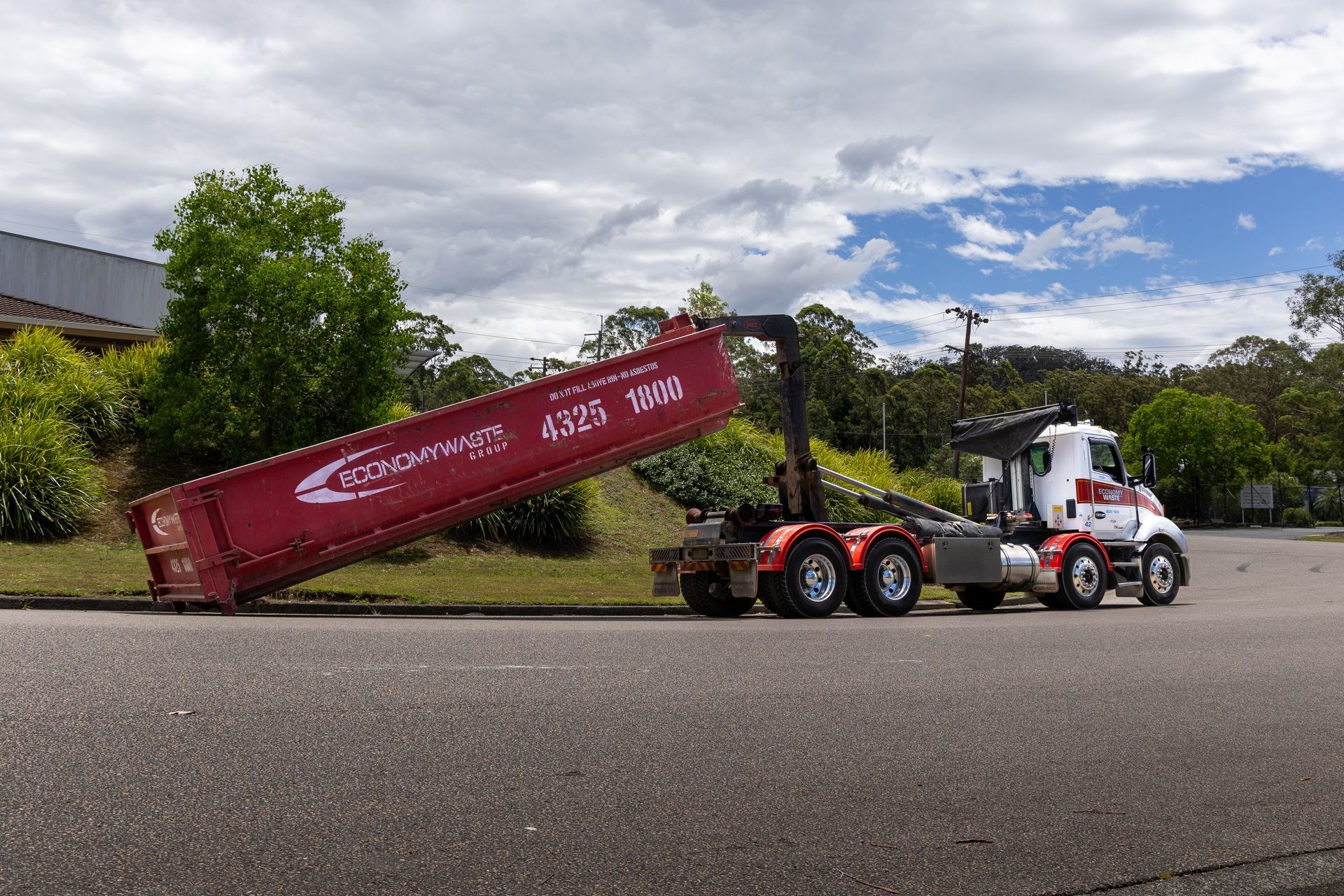 Red truck lifting a red dumpster on asphalt road — Economy Waste Group | Skip Bins Central Coast