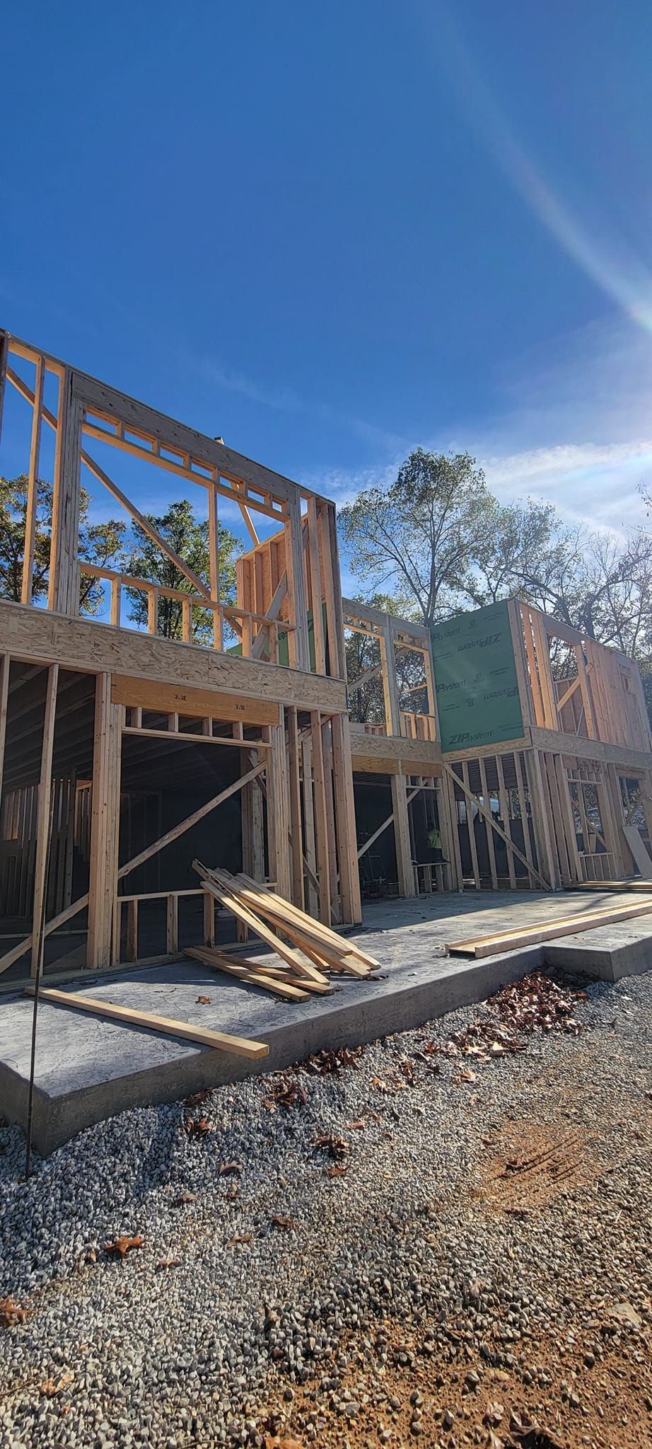 Wooden house frame under construction against a blue sky, surrounded by gravel and bare earth.
