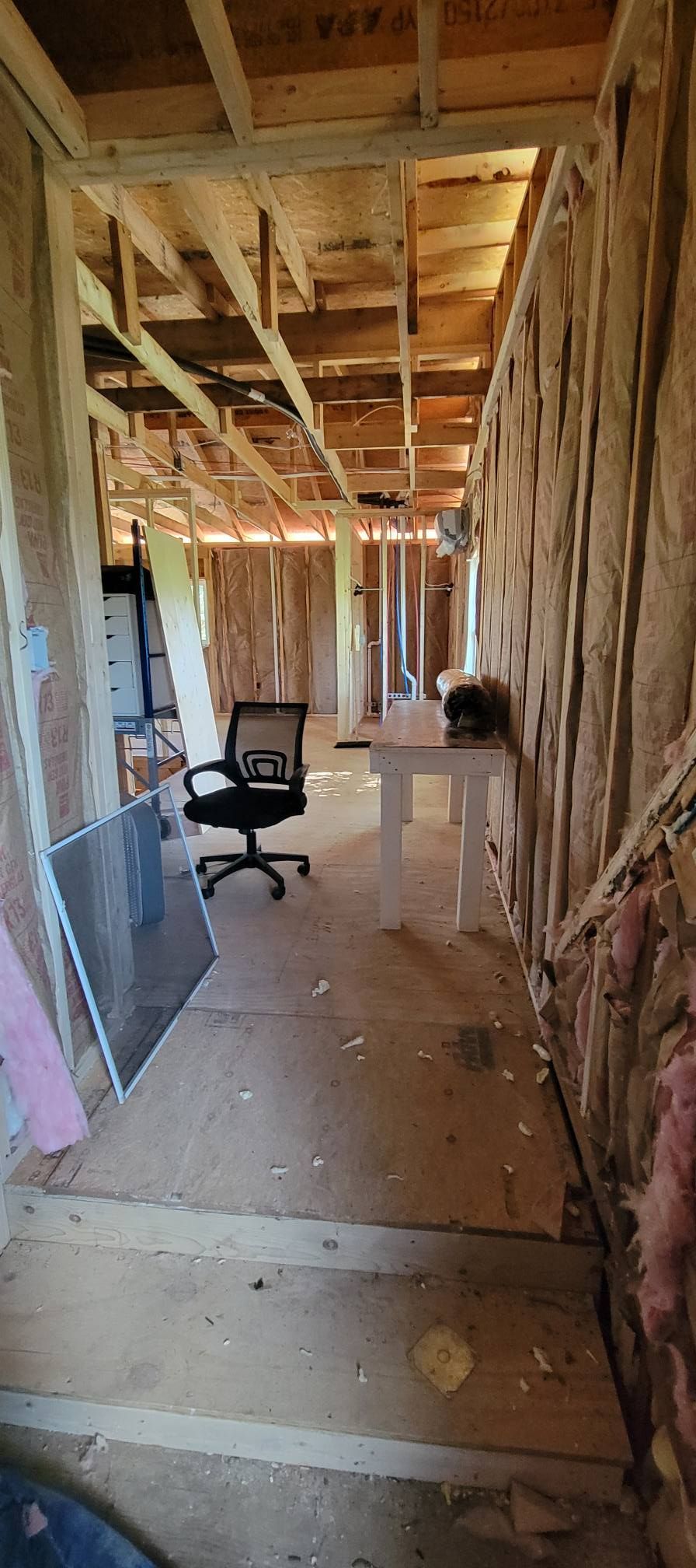 Interior of a room under construction, with exposed wood framing, insulation, and a desk chair.