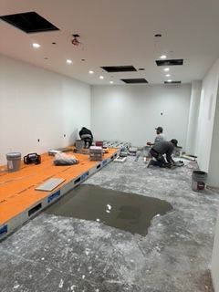 Construction workers installing tile flooring in a room with white walls, lighting, and cut-out ceiling panels.