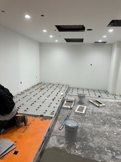 Room under construction; tile installation in progress. Worker, grey bucket, mortar, tile spacers, and white walls.