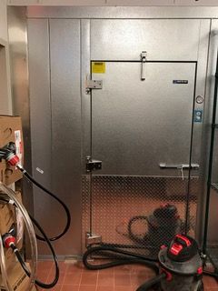 Metal walk-in refrigerator door, with a latch. Red tile floor. Black hose and vacuum cleaner in foreground.