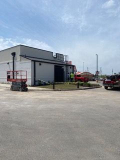 Exterior of a building with construction work. A lift, workers, and trucks are visible on a paved lot under a cloudy sky.