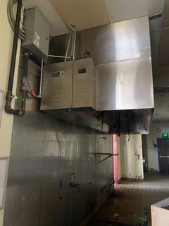 Stainless steel kitchen hood over a cooking area with metal boxes, piping, and conduit on the wall.