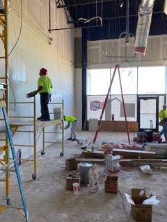 Construction workers drywalling interior walls; scaffold, tools, and materials on the floor.