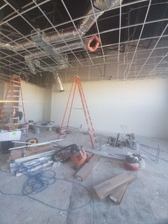 Construction site interior with exposed ceiling grid, ladders, and tools, walls are white.