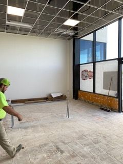 Construction in progress: Worker in a yellow shirt walks near a large window, open space with exposed floor, and ceiling grid.