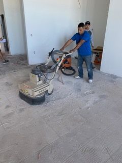 Man using floor grinder in an unfinished room. Another man watches. White walls, concrete floor.
