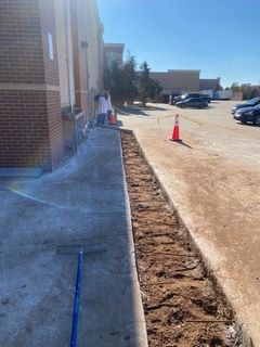 Sidewalk construction outside a brick building; soil trench, concrete, safety cone. Person in distance.