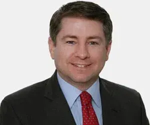 Studio portrait of a man in a dark suit, light blue shirt, and red tie, smiling against a white background