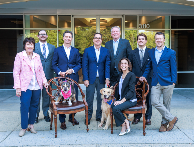 Eight people and two dogs posing outside a building entrance, smiling in business attire