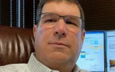 Close-up of a man wearing glasses in an office, seated by a computer screen and leather chair.