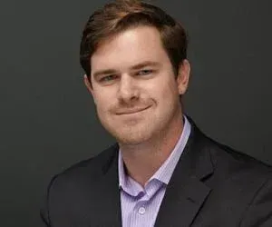 Portrait of a man in a dark blazer and light purple shirt, smiling against a gray background