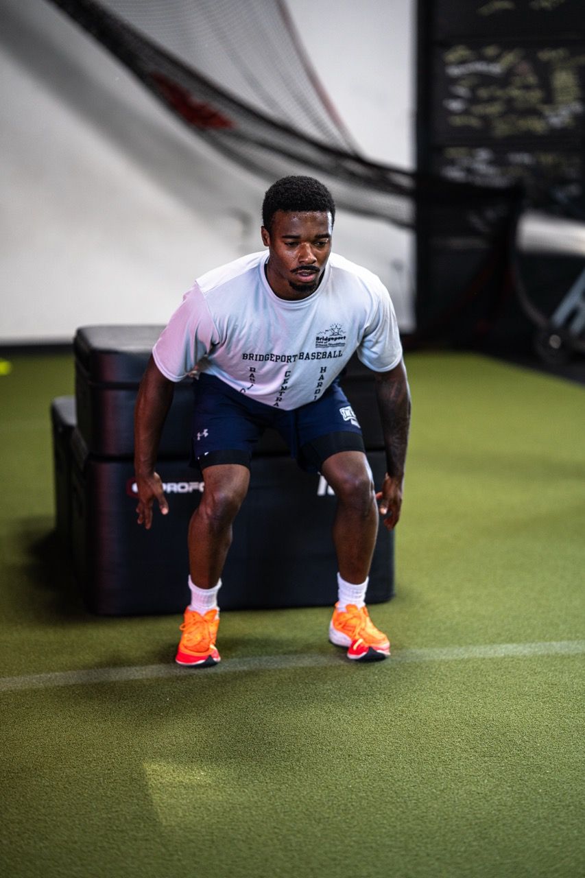 Athlete in a low squat stance on indoor turf at Division X Training in Branford, CT, wearing a white shirt, dark shorts, and bright orange shoes.