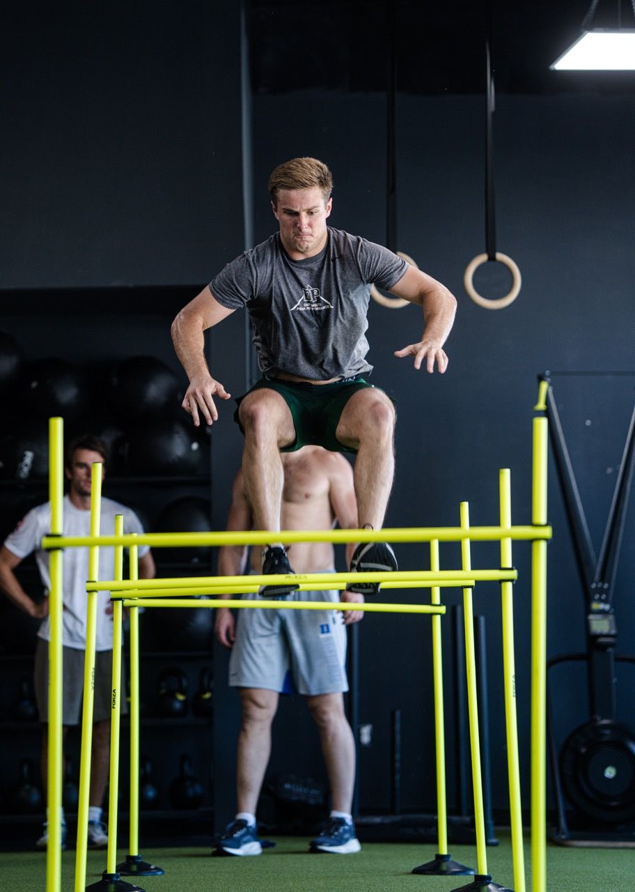 Man jumping over yellow hurdles with another person spotting in a gym, ring hanging behind