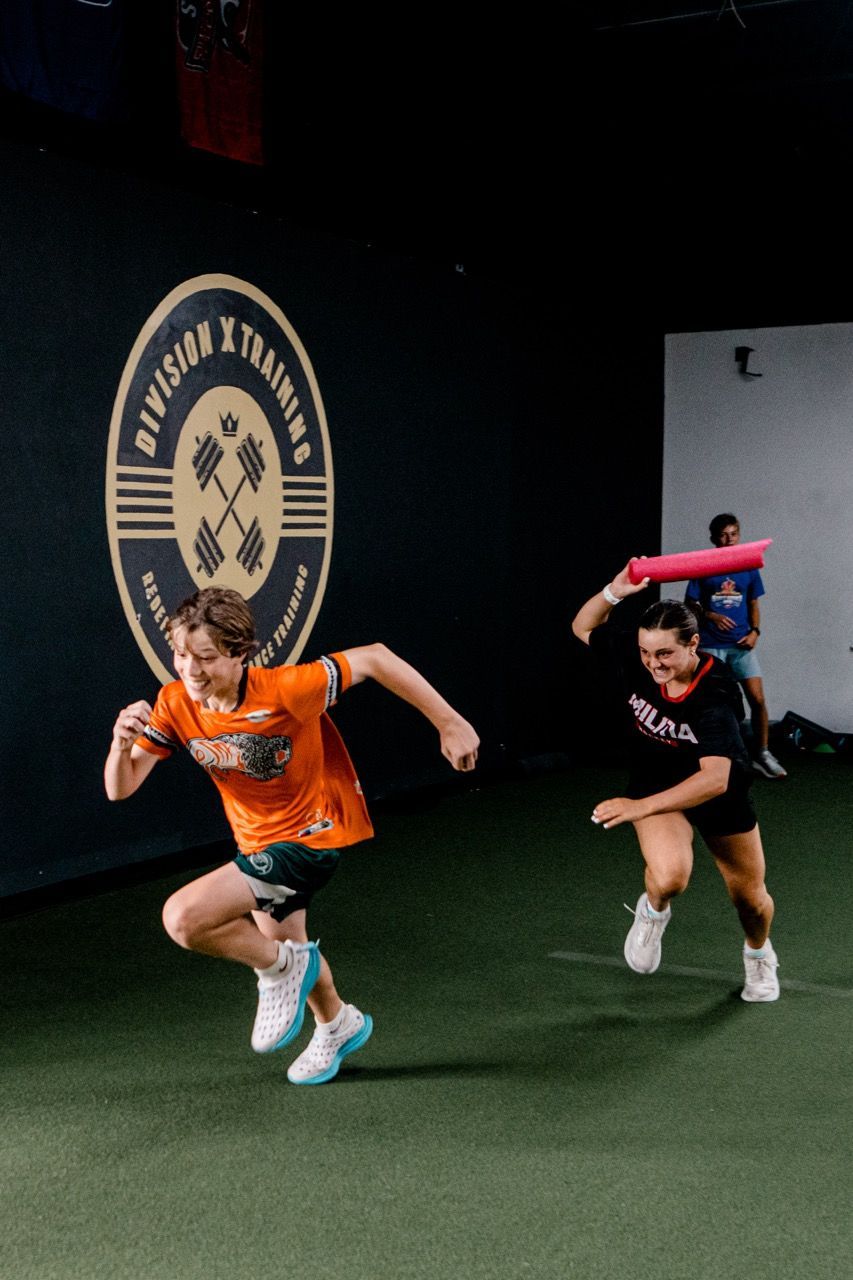 Two athletes sprint in Division X Training in Branford, CT, one in an orange shirt and one in black holding a pink bar overhead.
