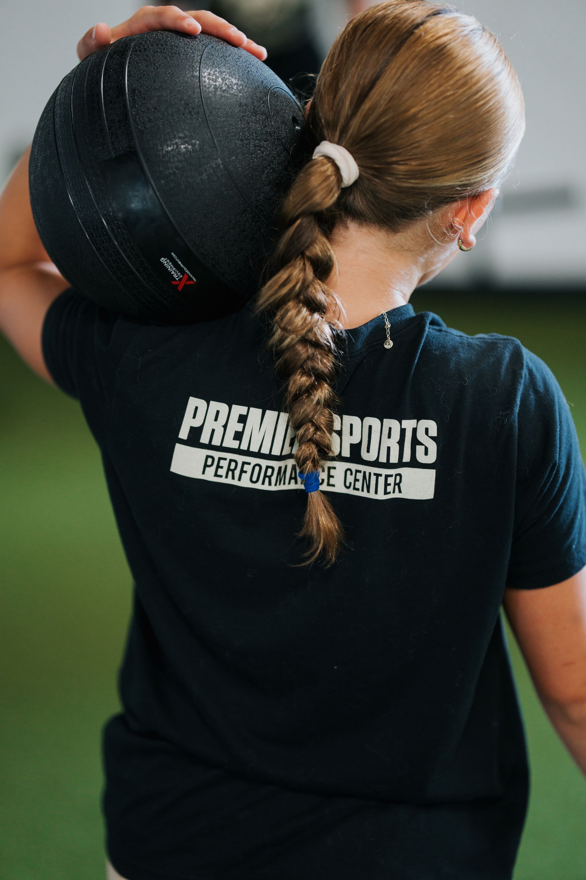 Athlete with braided hair carrying a black medicine ball on her shoulder, wearing a black sports shirt.