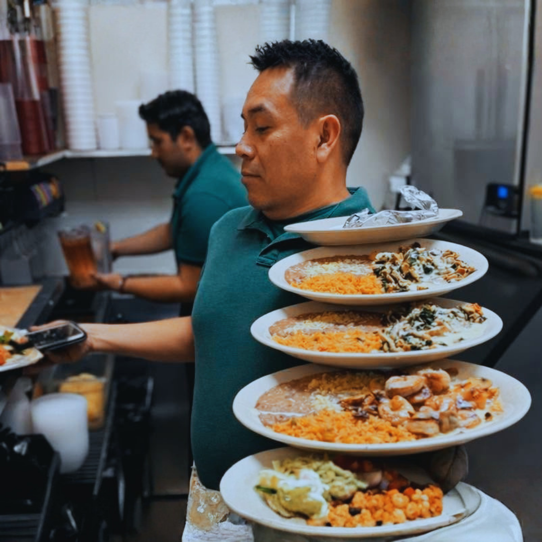 Waiter holding a stack of plated food in a busy kitchen, with another worker in the background
