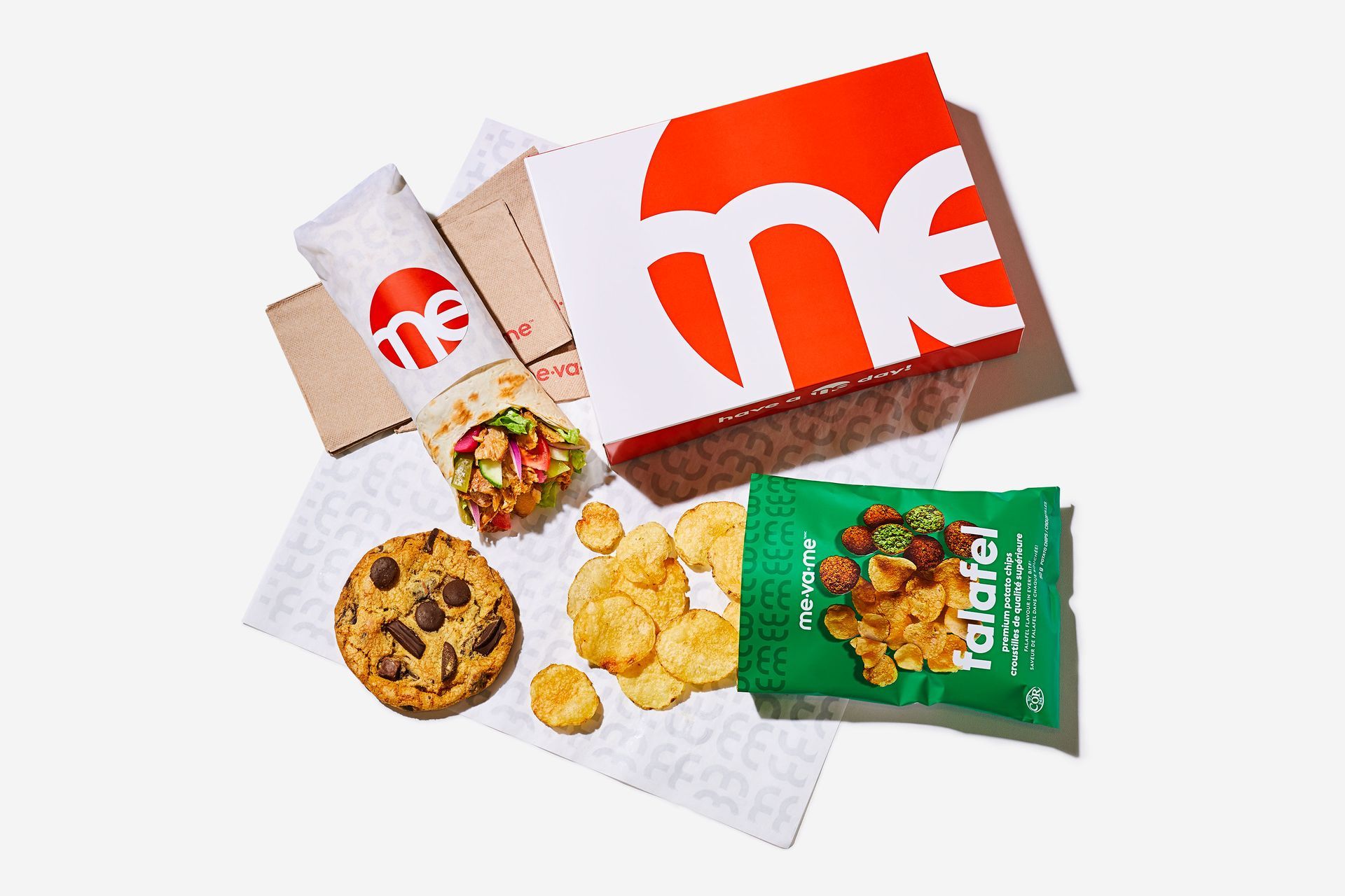 A box of food with a sandwich , cookie , chips and a bag of falafel.